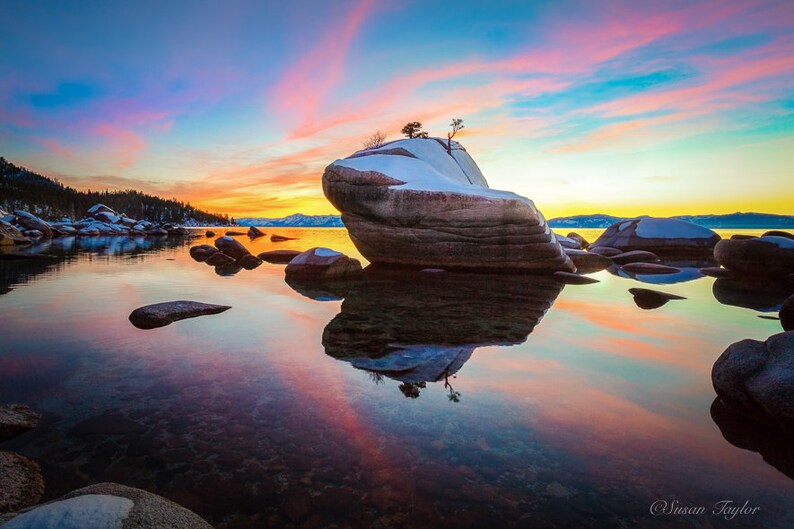 Puede incluir: Una gran formaci&oacute;n rocosa con un peque&ntilde;o &aacute;rbol creciendo en la cima, cubierta de nieve, se encuentra en un lago tranquilo al atardecer. El agua refleja el cielo colorido, creando una hermosa escena.