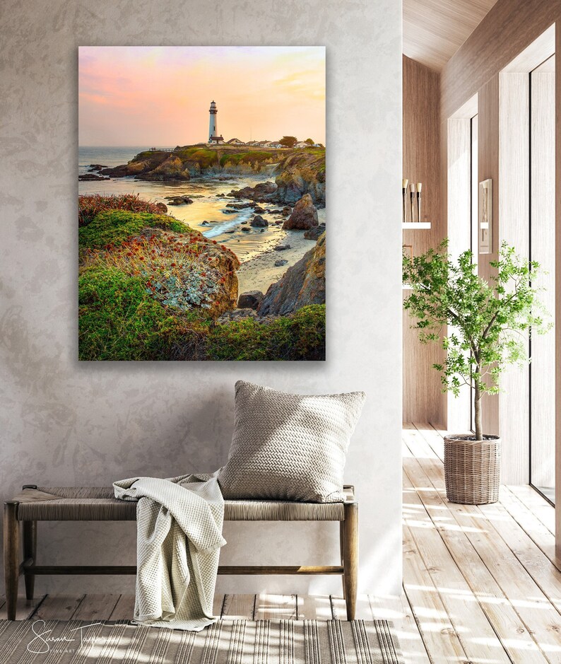 May include: A white lighthouse stands tall on a rocky cliff overlooking a calm ocean at sunset. The sky is a soft pink and orange, and the water is a deep blue. The lighthouse is surrounded by green vegetation and rocks.
