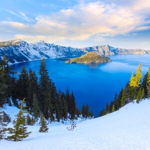May include: A panoramic view of Crater Lake in Oregon, with snow-capped mountains and a deep blue lake. The lake is surrounded by evergreen trees and a small island in the center.