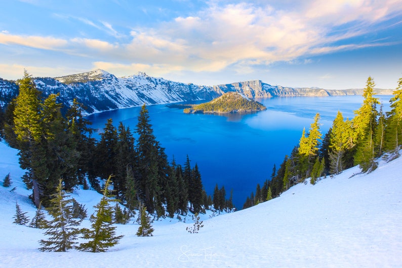 May include: A panoramic view of Crater Lake in Oregon, with snow-capped mountains and a deep blue lake. The lake is surrounded by evergreen trees and a small island in the center.