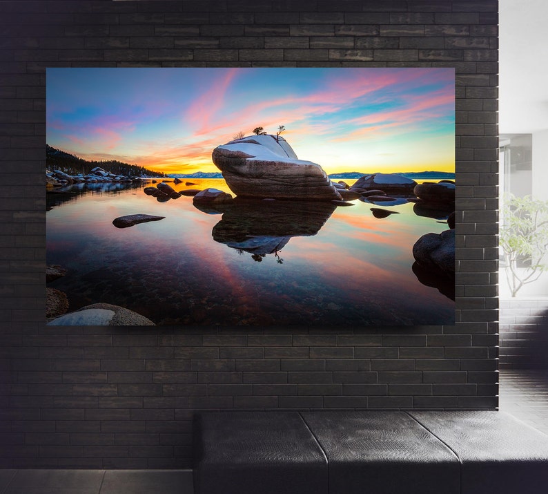 May include: A scenic photograph of a large rock formation with a small tree on top, surrounded by water and smaller rocks. The water reflects the colorful sky, which is a mix of pink, orange, and blue. The scene is calm and peaceful, with a soft, natural light.