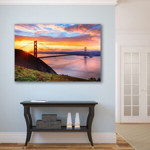 K&ouml;nnte beinhalten: Ein Foto der Golden Gate Bridge in San Francisco, Kalifornien. Die Br&uuml;cke wird vom goldenen Licht des Sonnenaufgangs beleuchtet, mit einem bew&ouml;lkten Himmel im Hintergrund. Die Br&uuml;cke ist aus der Ferne zu sehen, mit dem Wasser der Bucht im Vordergrund.