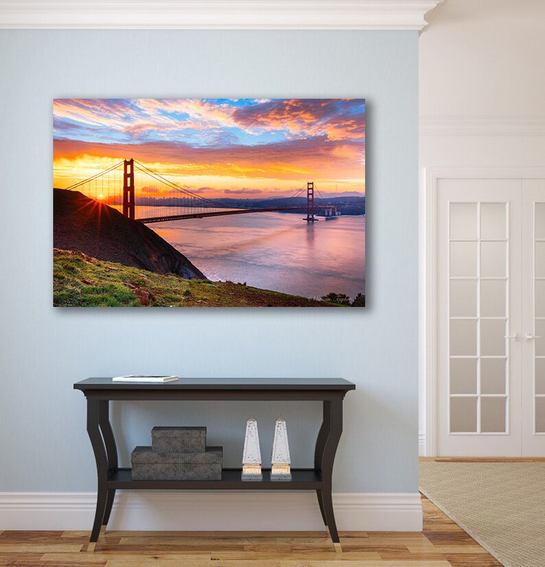 K&ouml;nnte beinhalten: Ein Foto der Golden Gate Bridge in San Francisco, Kalifornien. Die Br&uuml;cke wird vom goldenen Licht des Sonnenaufgangs beleuchtet, mit einem bew&ouml;lkten Himmel im Hintergrund. Die Br&uuml;cke ist aus der Ferne zu sehen, mit dem Wasser der Bucht im Vordergrund.