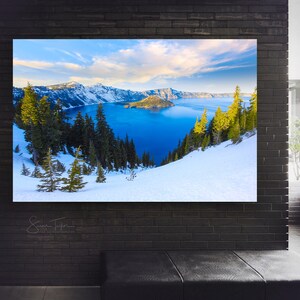 May include: A panoramic view of Crater Lake in Oregon, with snow-covered hills and a blue lake surrounded by forested mountains. The sky is a bright blue with white clouds.