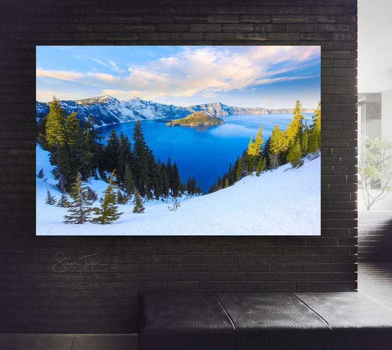 May include: A panoramic view of Crater Lake in Oregon, with snow-covered hills and a blue lake surrounded by forested mountains. The sky is a bright blue with white clouds.