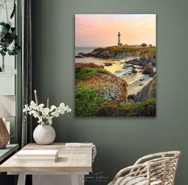 May include: A white lighthouse stands on a rocky cliff overlooking a calm ocean at sunset. The sky is a soft pink and orange, and the water is a deep blue. The lighthouse is surrounded by green grass and rocks.