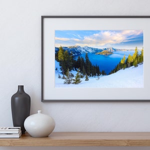 May include: Framed photograph of a snowy mountain landscape with a blue lake in the distance. The lake is surrounded by snow-covered mountains and trees. The sky is blue with white clouds.