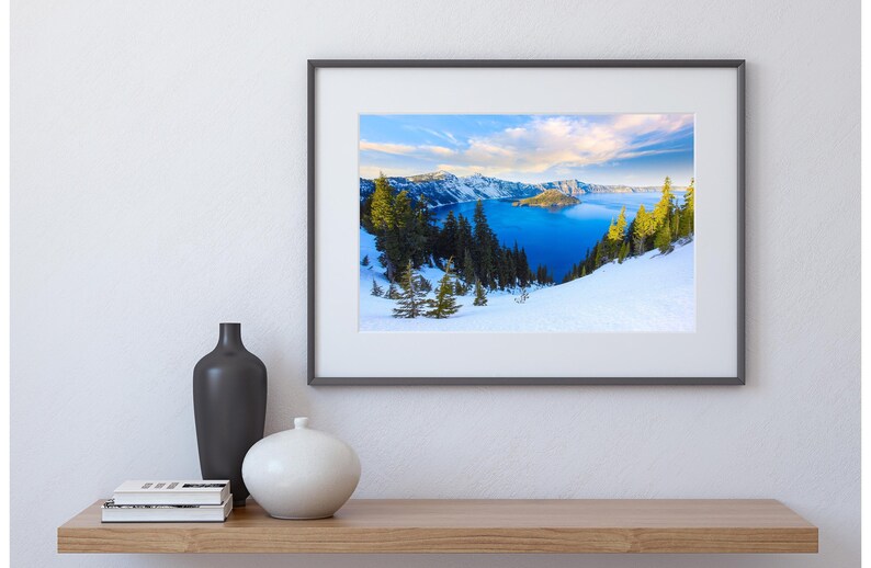 May include: Framed photograph of a snowy mountain landscape with a blue lake in the distance. The lake is surrounded by snow-covered mountains and trees. The sky is blue with white clouds.