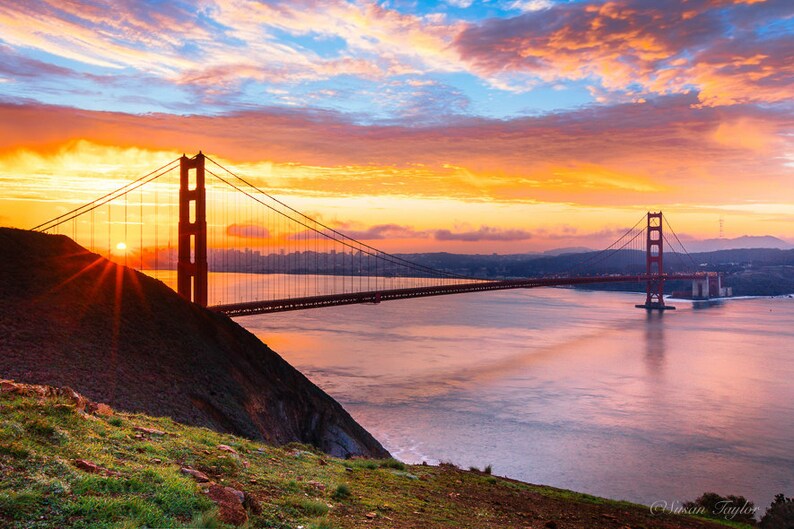 K&ouml;nnte beinhalten: Eine malerische Aussicht auf die Golden Gate Bridge in San Francisco, Kalifornien. Die Br&uuml;cke ist vor einem leuchtend orangefarbenen und rosafarbenen Sonnenaufganghimmel silhouettiert. Die Sonne scheint hell hinter der Br&uuml;cke und erzeugt einen Sterneneffekt.