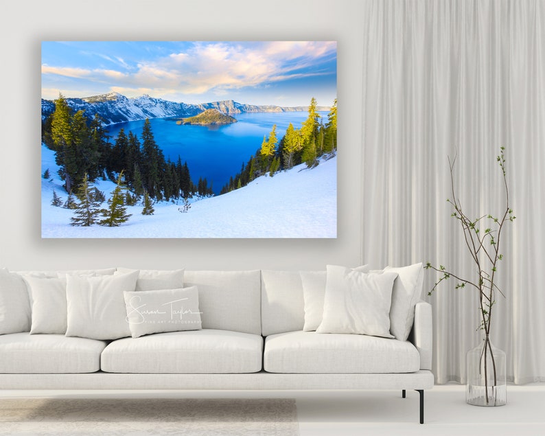 May include: A panoramic view of Crater Lake in Oregon, with snow-covered hills and a blue lake in the background. The lake is surrounded by trees and mountains. The sky is blue with white clouds.