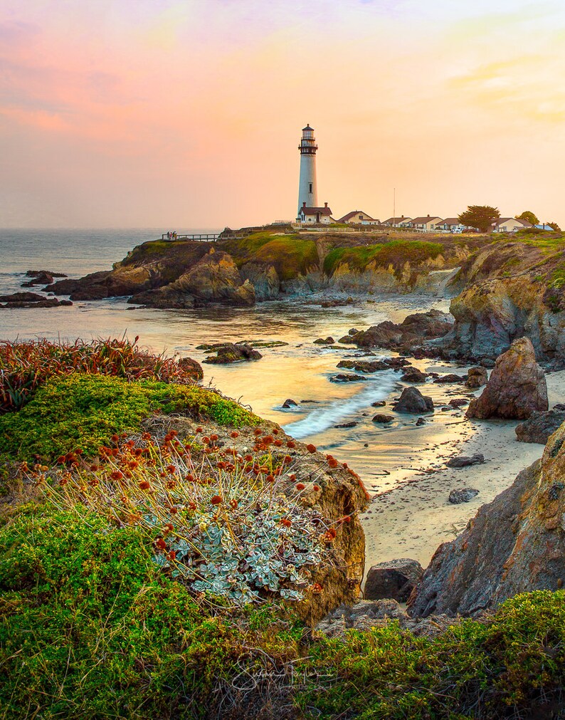 May include: A white lighthouse stands on a rocky cliff overlooking a calm ocean at sunset. The sky is a soft pink and purple, and the water is a shimmering gold.