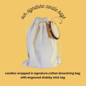 May include: A natural canvas drawstring bag with a wooden tag. The bag is labeled "our signature candle bags" and contains candles. The tag reads "candles wrapped in signature cotton drawstring bag with engraved shabby wick tag."