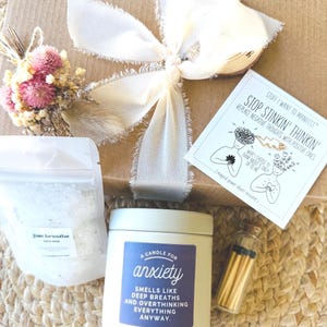 May include: A gift box with a white ribbon, dried flowers, and a candle labeled "A Candle for Anxiety." The box also contains bath salts, a small vial of matches, and a card with the text "Stop Stinkin' Thinkin'."