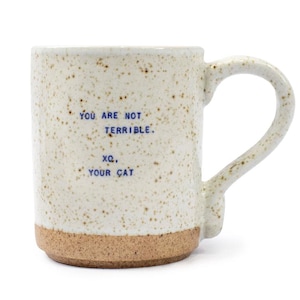May include: Cream-colored ceramic mug with a speckled finish and a brown base. The mug features the text "YOU ARE NOT TERRIBLE. XO, YOUR CAT" in blue. The handle is a matching cream color.
