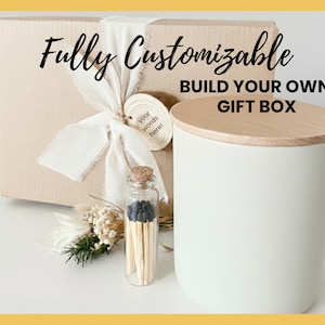 May include: A white cylindrical candle with a wooden lid sits next to a gift box tied with a cream ribbon. A small glass bottle filled with matches and a decorative floral arrangement are also present. The text reads "Fully Customizable, Build Your Own Gift Box."