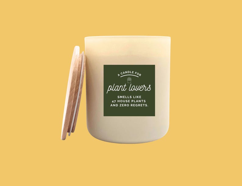 May include: A cream-colored candle with a wooden lid, set against a yellow background. The candle has a green label that reads "A candle for plant lovers" and "Smells like 47 house plants and zero regrets."