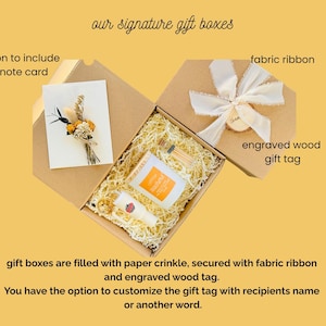 May include: Open gift boxes with paper crinkle, a note card with dried flowers, and small products. The boxes are secured with fabric ribbon and an engraved wood gift tag. Text reads "our signature gift boxes".