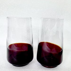 Könnte beinhalten: Zwei klare, zylindrische Gläser, die jeweils eine dunkelrote Flüssigkeit enthalten. Die Gläser sind leicht geneigt, und die Flüssigkeit füllt den unteren Teil jedes Glases. Der Hintergrund ist eine schlichte weiße Oberfläche.