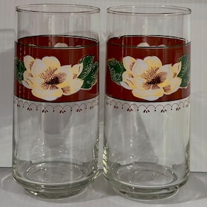 Puede incluir: Dos vasos de beber de vidrio transparente con una banda roja y un diseño de flor de magnolia blanca. La banda tiene un borde festoneado con puntos rojos.