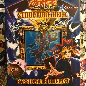 Puede incluir: Una caja de mazo de estructura de juego de cartas coleccionables Yu-Gi-Oh! azul y dorada con el texto "Legendary Duelist Structure Deck: Passionate Duelist" y una imagen de un personaje de dibujos animados sosteniendo una carta.