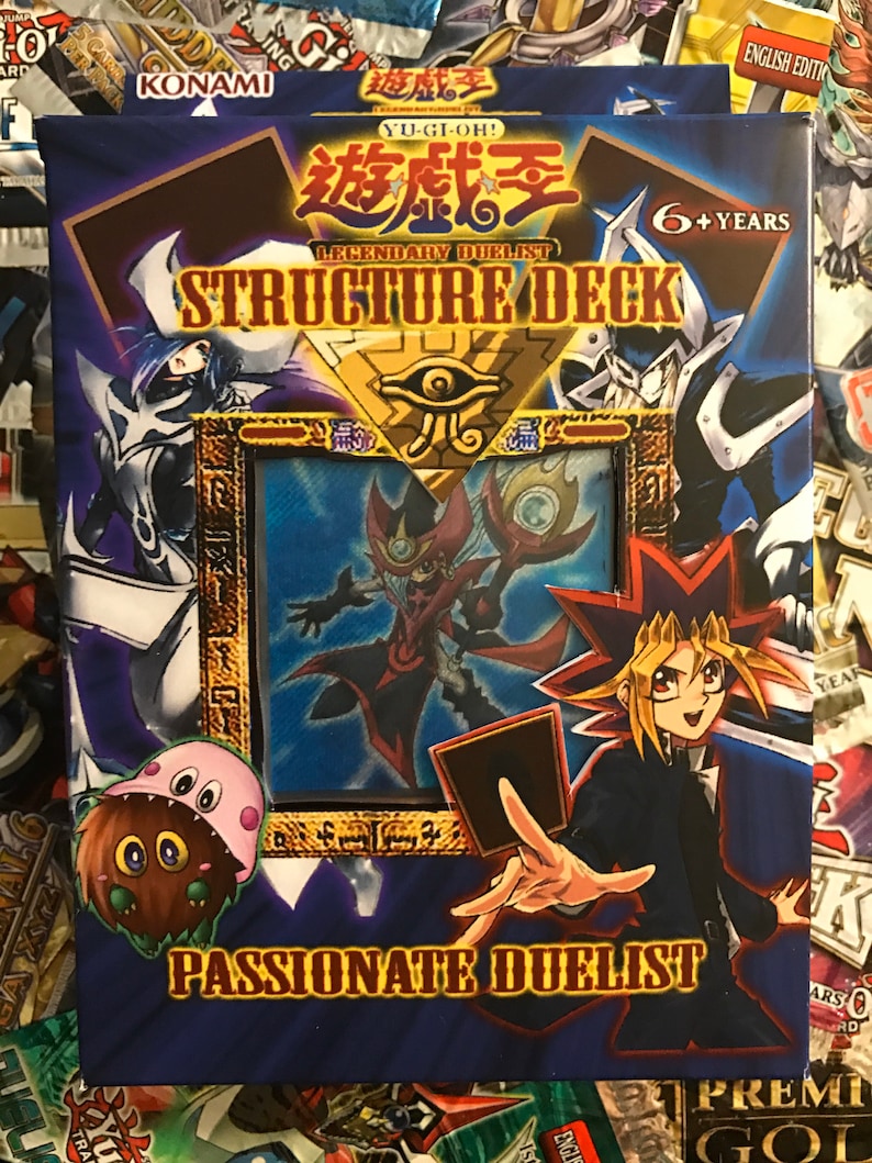 Puede incluir: Una caja de mazo de estructura de juego de cartas coleccionables Yu-Gi-Oh! azul y dorada con el texto "Legendary Duelist Structure Deck: Passionate Duelist" y una imagen de un personaje de dibujos animados sosteniendo una carta.