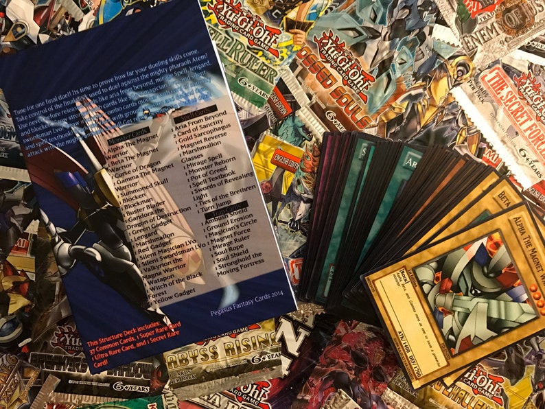 Puede incluir: Una colecci&oacute;n de cartas de intercambio de Yu-Gi-Oh!, que incluye un paquete de refuerzo, una caja de mazo y un conjunto de cartas con el monstruo Alpha the Magnet Warrior. Las cartas est&aacute;n dispuestas sobre una mesa con otros productos de Yu-Gi-Oh! en el fondo.