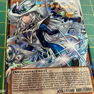 Pode incluir: Uma carta colecionável holográfica de Yu-Gi-Oh! intitulada Silent Magician, the Silent Fighter. A carta mostra um personagem de cabelos brancos com chapéu branco e traje azul, com o texto Spellcaster/Effect e ATK/DEF 3500/1000.