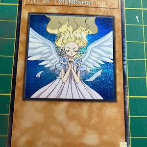 Op de afbeelding: Een rechthoekige ruilkaart met een beige rand en een holografische blauwe achtergrond. De centrale illustratie toont een gevleugeld figuur met blond haar en een witte jurk, met gevouwen handen. De tekst "Ties of Friendship" staat bovenaan gedrukt.
