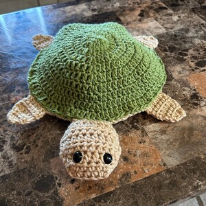 May include: A handmade crochet turtle in shades of green and tan. The turtle has a green shell, tan flippers, and a tan head with black eyes. The turtle is lying on a brown surface.