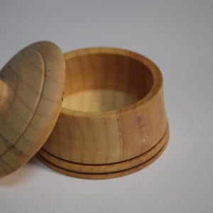Puede incluir: Un recipiente peque&ntilde;o y redondo de madera clara con una tapa a juego. El recipiente presenta un acabado de madera natural lisa y dos l&iacute;neas horizontales finas y oscuras grabadas alrededor de la base. La tapa tiene un pomo central.