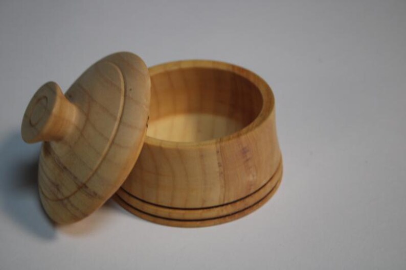Puede incluir: Un recipiente peque&ntilde;o y redondo de madera clara con una tapa a juego. El recipiente presenta un acabado de madera natural lisa y dos l&iacute;neas horizontales finas y oscuras grabadas alrededor de la base. La tapa tiene un pomo central.
