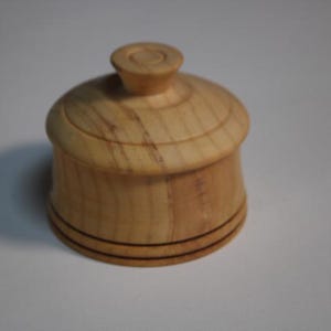 Puede incluir: Un peque&ntilde;o recipiente redondo de madera de color claro con una tapa a juego. La tapa cuenta con un pomo central y la base del recipiente est&aacute; decorada con dos finas l&iacute;neas horizontales oscuras grabadas alrededor del contorno.