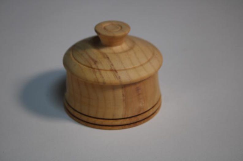 Puede incluir: Un peque&ntilde;o recipiente redondo de madera de color claro con una tapa a juego. La tapa cuenta con un pomo central y la base del recipiente est&aacute; decorada con dos finas l&iacute;neas horizontales oscuras grabadas alrededor del contorno.
