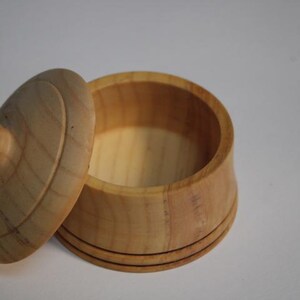 Puede incluir: Un peque&ntilde;o recipiente redondo de madera de color claro con su tapa a juego colocada al lado. El recipiente presenta un acabado liso con veta de madera natural y un pomo circular en el centro de la tapa. El dise&ntilde;o es sencillo y de base cil&iacute;ndrica.