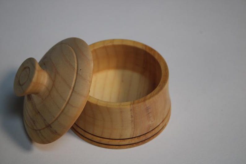 Puede incluir: Un peque&ntilde;o recipiente redondo de madera de color claro con su tapa a juego colocada al lado. El recipiente presenta un acabado liso con veta de madera natural y un pomo circular en el centro de la tapa. El dise&ntilde;o es sencillo y de base cil&iacute;ndrica.