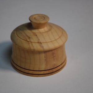 Puede incluir: Un peque&ntilde;o recipiente redondo de madera de color claro con una tapa a juego. La tapa cuenta con un pomo central. El cuerpo del recipiente tiene un acabado suave y pulido con patrones de veta de madera natural visibles y dos l&iacute;neas oscuras.