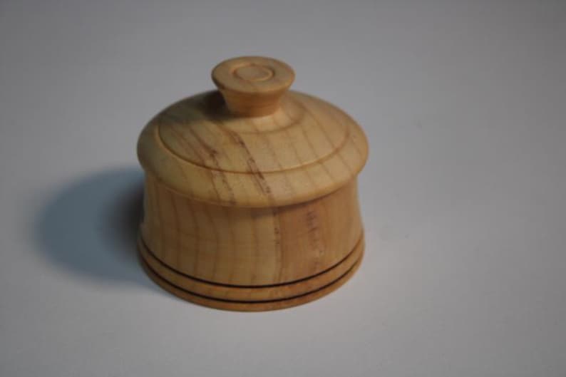 Puede incluir: Un peque&ntilde;o recipiente redondo de madera de color claro con una tapa a juego. La tapa cuenta con un pomo central. El cuerpo del recipiente tiene un acabado suave y pulido con patrones de veta de madera natural visibles y dos l&iacute;neas oscuras.