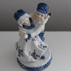May include: A porcelain figurine of a couple dancing. The woman is wearing a white dress with blue and gold floral designs. The man is wearing blue trousers and a blue hat. The figurine is on a white base with blue and gold designs.