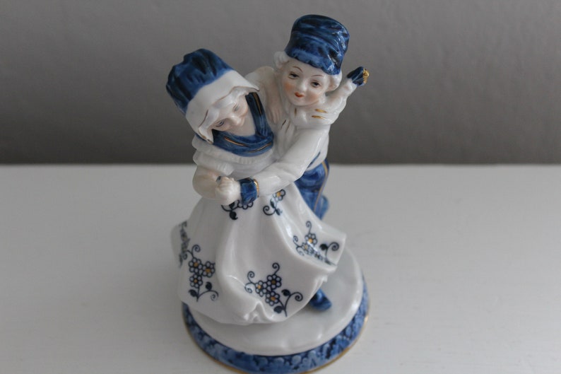 May include: A porcelain figurine of a couple dancing. The woman is wearing a white dress with blue and gold floral designs. The man is wearing blue trousers and a blue hat. The figurine is on a white base with blue and gold designs.