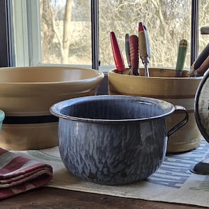 May include: A collection of vintage kitchenware, including a blue and gray enamelware chamber pot with a handle, yellow and blue striped bowls, and a scale. Various kitchen utensils are in a yellow and brown container.