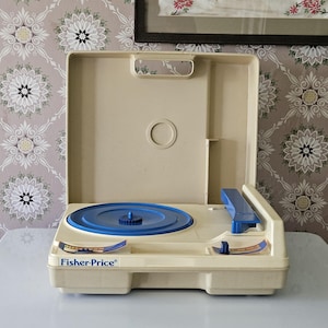 May include: A beige plastic Fisher-Price toy record player with a blue circular turntable and a blue tone arm. The device features a hinged lid, a handle, and control knobs with blue and orange accents. The Fisher-Price logo is printed in blue.