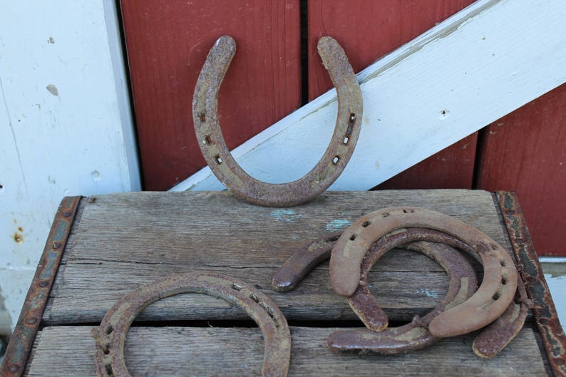 May include: Five rusty horseshoes, one is hanging on a white and red wooden door, the other four are on a wooden surface.