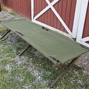 May include: A portable olive green canvas cot featuring a sturdy, foldable metal frame with X-shaped legs. The rectangular sleeping surface is stretched taut across the horizontal support bars, providing a flat, elevated platform for resting.