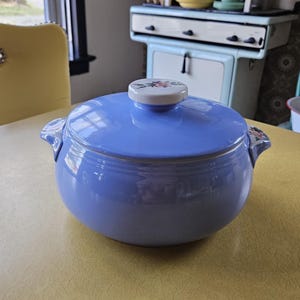May include: A light blue ceramic casserole dish with a lid. The lid has a white knob with a floral design. The dish has two handles and is sitting on a yellow table. The dish is a vintage kitchenware item.