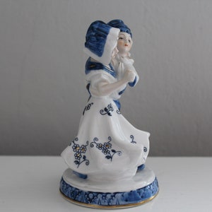 May include: A porcelain figurine of a couple in traditional Dutch clothing. The woman is wearing a blue and white dress with a blue hat, and the man is wearing a blue and white jacket with a blue hat. The figurine is painted in blue and white with gold accents.