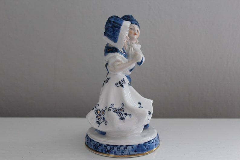 May include: A porcelain figurine of a couple in traditional Dutch clothing. The woman is wearing a blue and white dress with a blue hat, and the man is wearing a blue and white jacket with a blue hat. The figurine is painted in blue and white with gold accents.