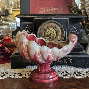 May include: A decorative, vintage ceramic bowl with a red and white marbled design. The bowl has a scalloped rim and sits on a pedestal base. Other antique items are in the background.