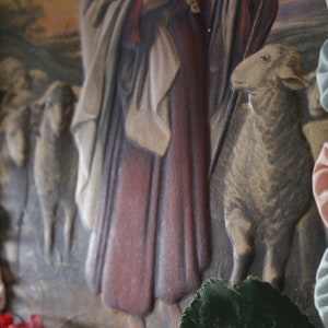 May include: A vintage religious image with a woman in a long red robe and a white shawl. Two sheep are in the foreground, one with its head turned to the side. The image is framed with a border of blue and pink fabric rosettes.