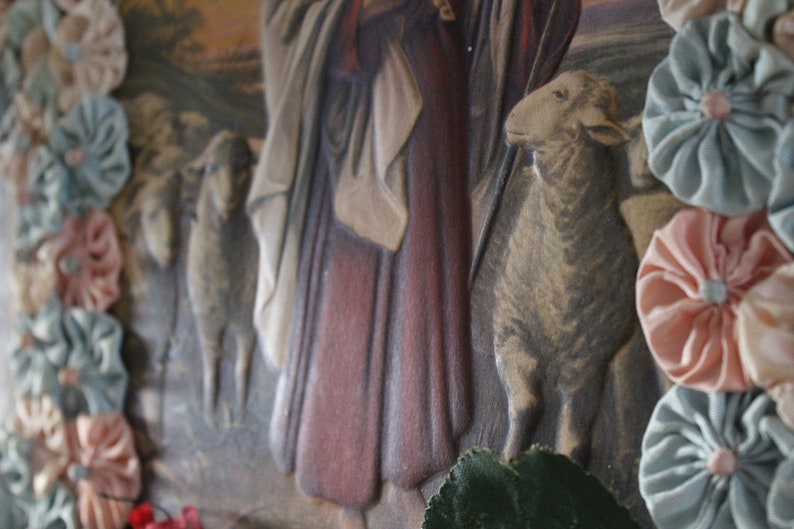 May include: A vintage religious image with a woman in a long red robe and a white shawl. Two sheep are in the foreground, one with its head turned to the side. The image is framed with a border of blue and pink fabric rosettes.