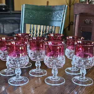 May include: A collection of nine vintage wine glasses with a clear, patterned base and a vibrant red rim. The glasses are arranged on a wooden table, showcasing their elegant design. The stemware is ideal for serving wine.